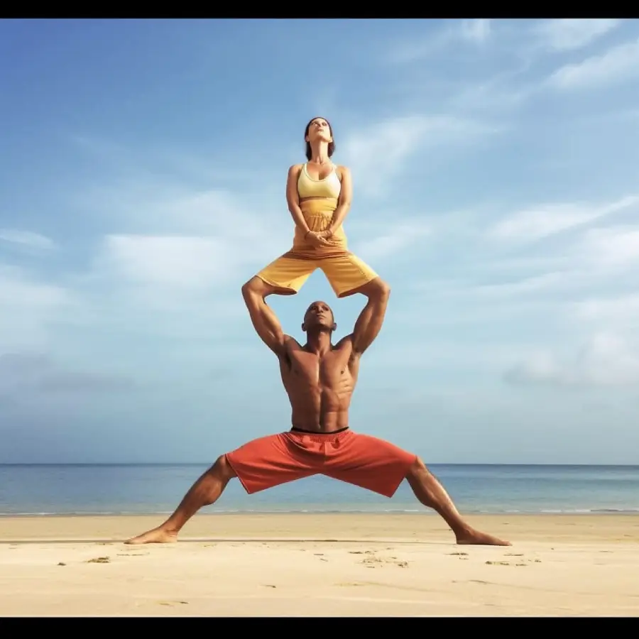 Two people perform an acro yoga pose on a beach. A man in red shorts stands with legs wide, holding a woman in yellow who balances on his shoulders against a blue sky and ocean backdrop.