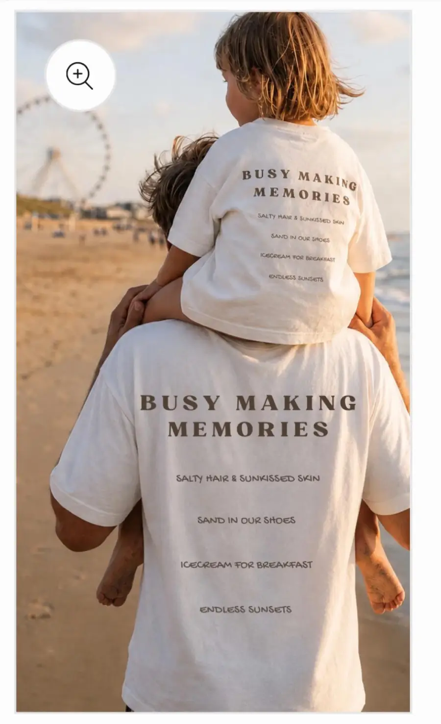 An adult carries a child on their shoulders at the beach. Both wear white shirts that say, “Busy Making Memories” with a list including “salty hair,” “sand in our shoes,” and “ice cream for breakfast.”