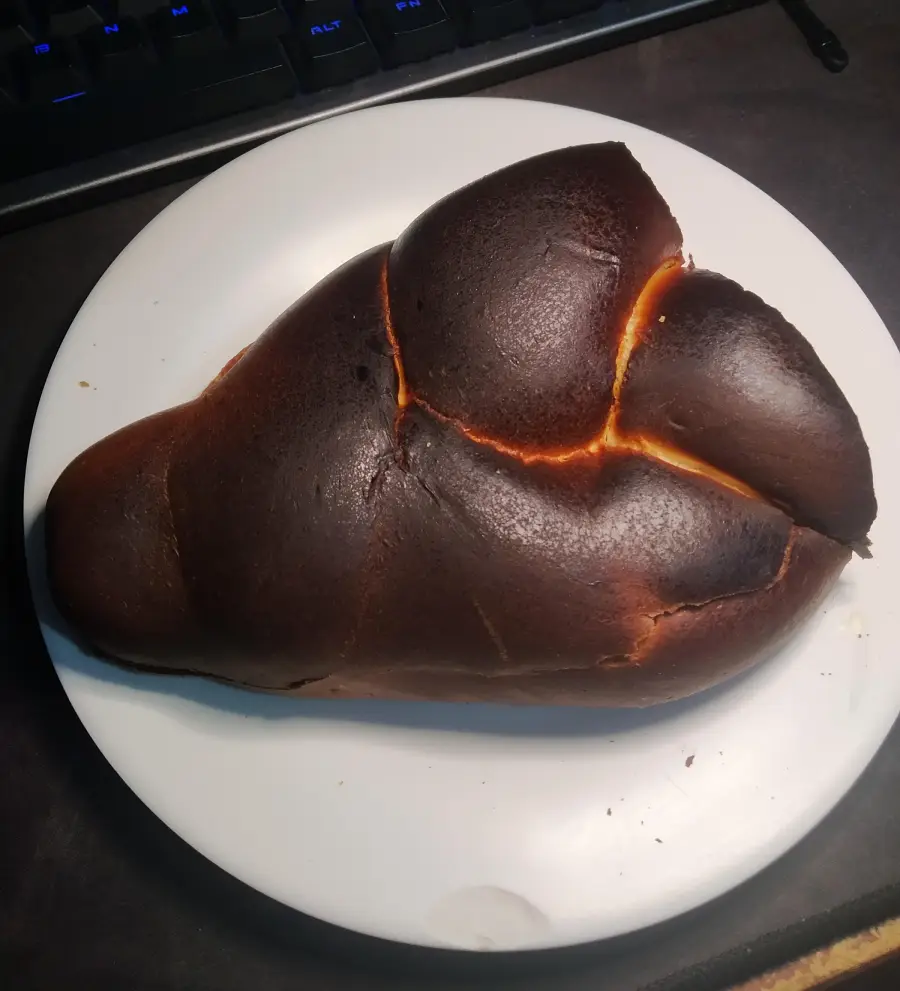 A loaf of bread with a very dark, almost burnt crust sits on a white plate, placed on a dark surface.