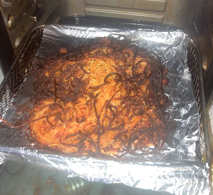 Burnt spaghetti sits on a foil-lined baking tray inside an oven. The noodles are blackened and stuck together, indicating they were overcooked or left in the oven for too long.