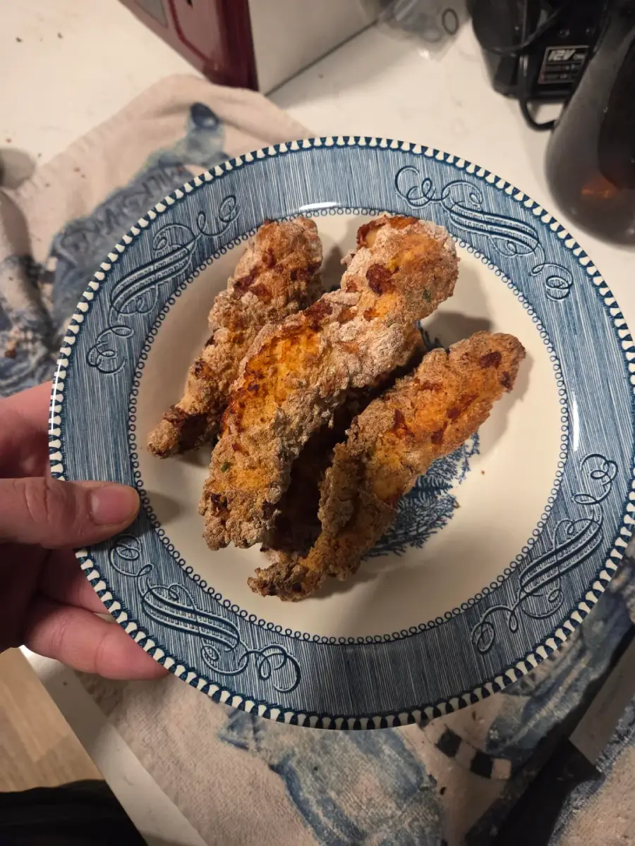 A hand holds a blue and white decorative plate with three pieces of golden-brown fried chicken tenders on it. The background shows a kitchen counter and patterned cloth.