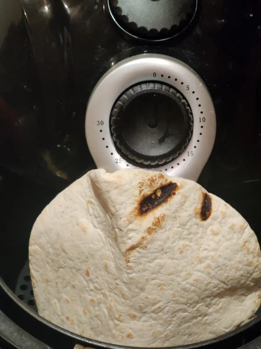 A flour tortilla with burnt spots rests inside an air fryer, positioned in front of the device’s timer dial set to about 25 minutes.