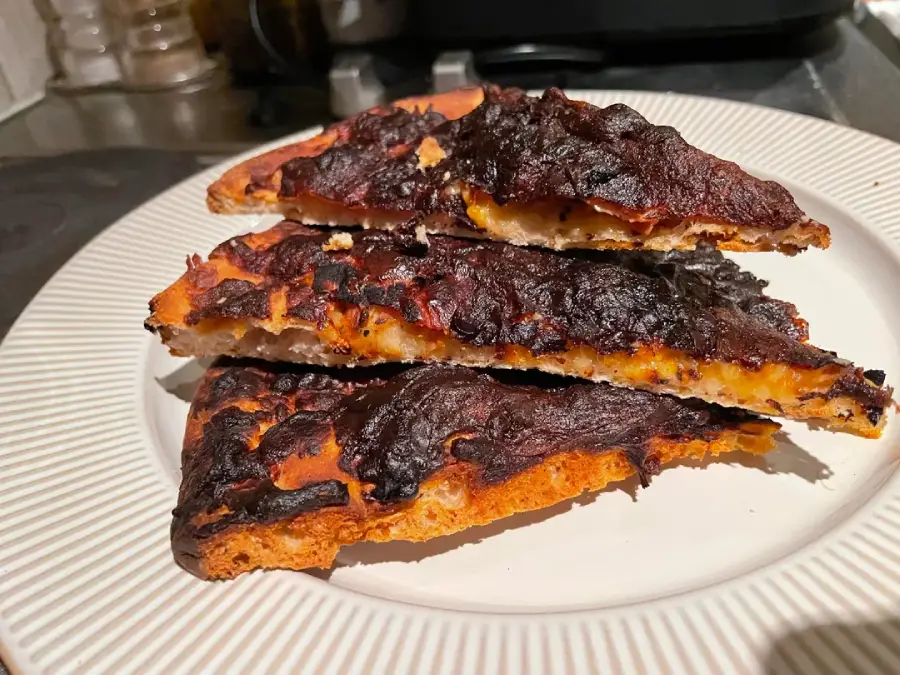 Three slices of burnt pizza stacked on a white plate, showing dark charred cheese and crust. The background includes kitchen items like salt and pepper shakers.
