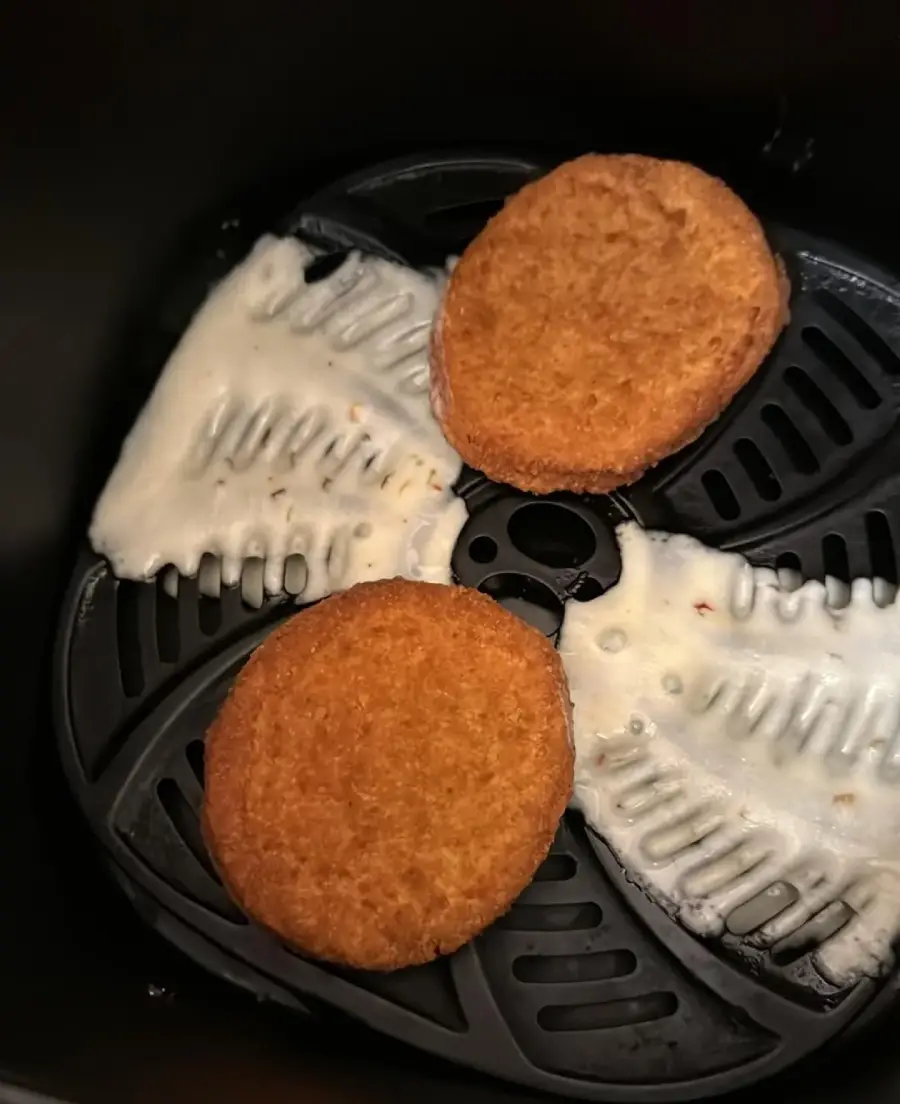 Two breaded patties sit on an air fryer tray with melted cheese stuck to the grate beneath them.