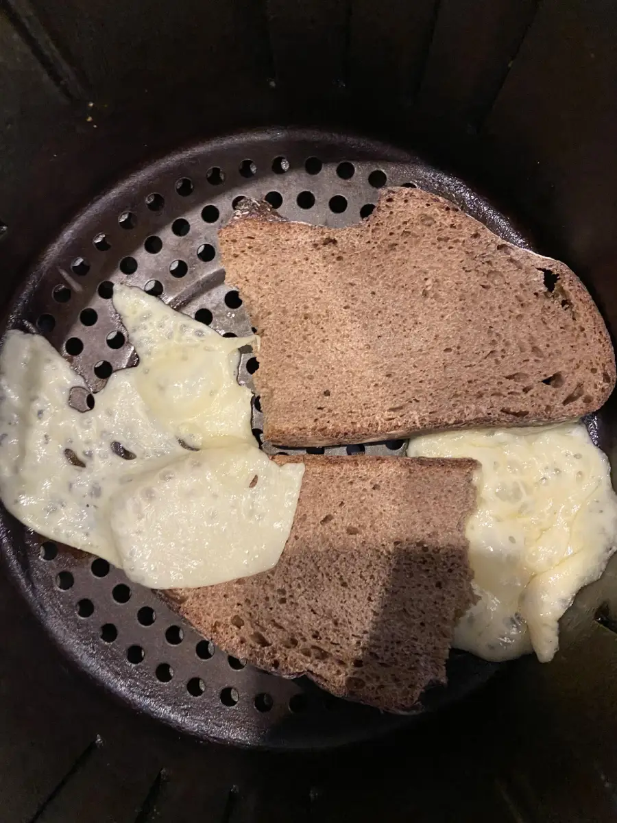 Two slices of dark bread and melted cheese sit on the perforated tray inside an air fryer. The cheese has spread unevenly beneath and beside the bread.