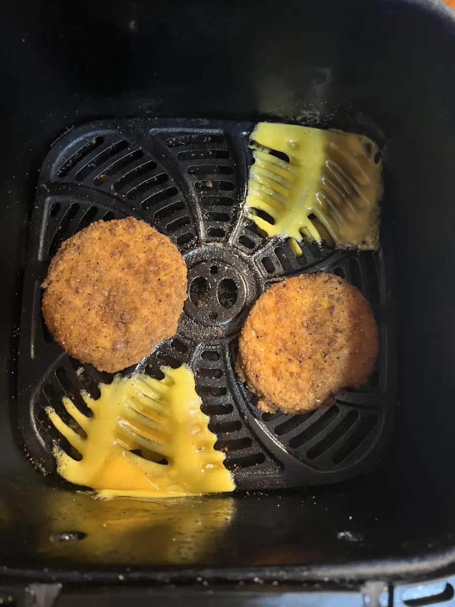 Two breaded patties and melted slices of cheese are inside an air fryer basket. The cheese has melted onto the bottom of the basket and the patties are spaced apart.