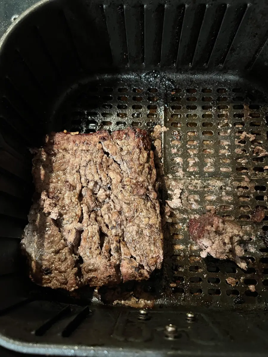 Cooked ground beef sits in the basket of an air fryer. The beef is unevenly shaped and browned, with some small pieces to the side. The air fryer basket has visible grill-like slots.