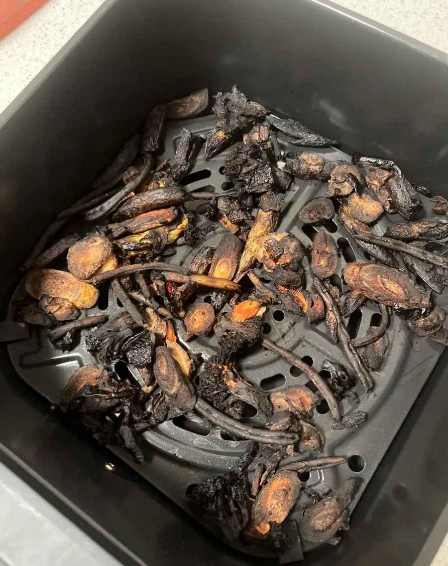 Burnt food scraps, possibly vegetables or mushrooms, are charred and blackened inside a black air fryer basket, indicating they have been overcooked.