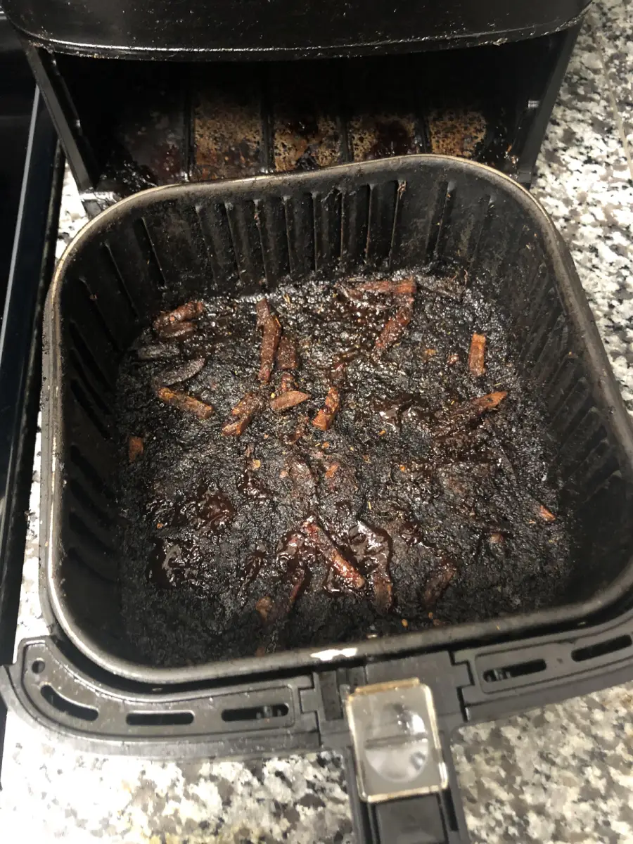Burnt food remnants and charred bits are stuck to the bottom of a black air fryer basket on a granite countertop, with the fryer drawer pulled out and some residue visible in the back.