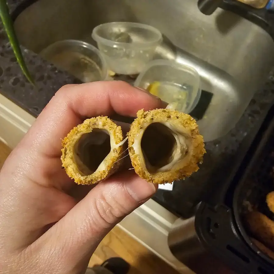 A hand holds two breaded mozzarella sticks with empty centers, showing melted cheese stuck to the inside but not filling the sticks. A kitchen sink with plastic containers is in the background.
