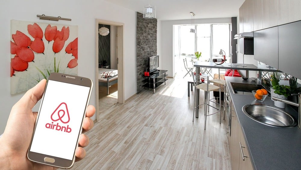 A hand holds a smartphone displaying the Airbnb logo in a modern, bright apartment with a kitchen, dining area, and a visible bedroom.