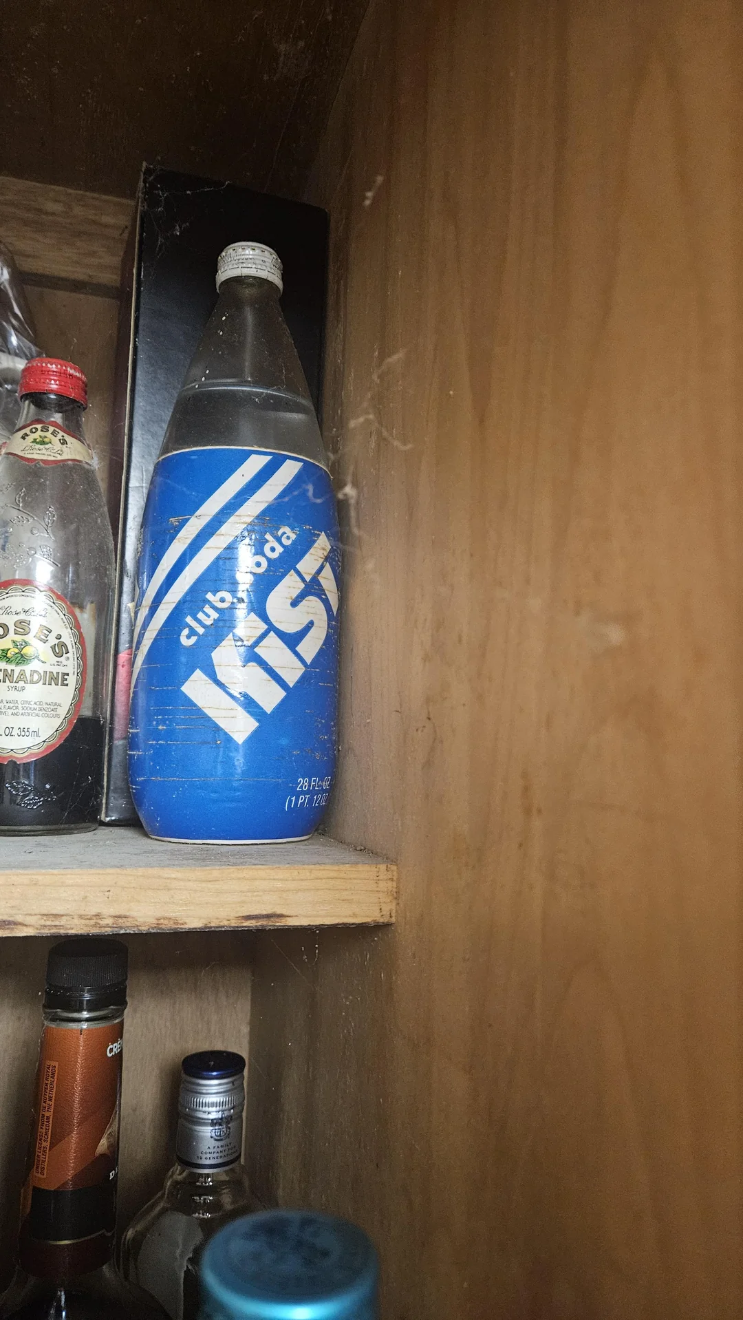 A dusty pantry shelf with a blue bottle labeled “Kist Club Soda” surrounded by other bottles, including a clear one on the left and some brown glass bottles below.