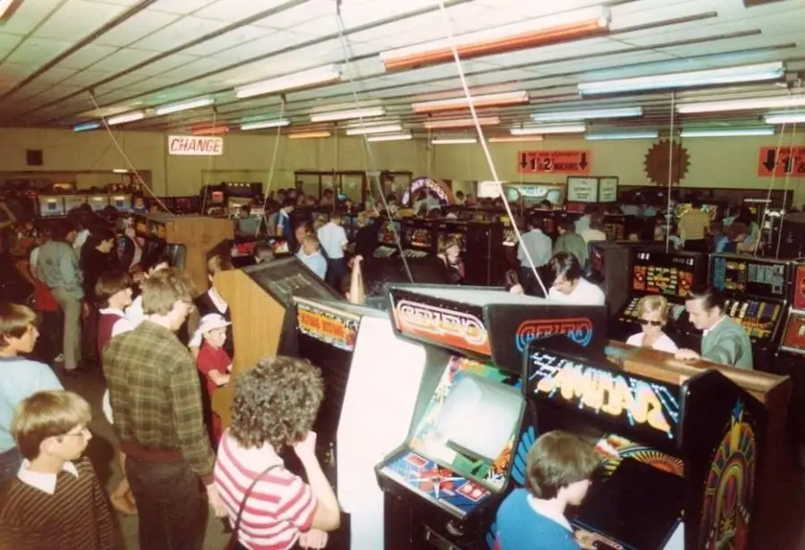 Crowded arcade filled with people playing vintage arcade machines; colorful signage and a lively atmosphere, with adults and children enjoying various games.