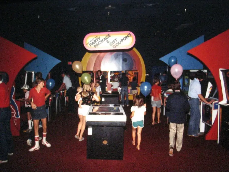 Children and adults play arcade games in a dimly lit room. Some hold colorful balloons. A large, colorful sign in the background reads “Party Package Gift Coupons.” The atmosphere appears lively and festive.
