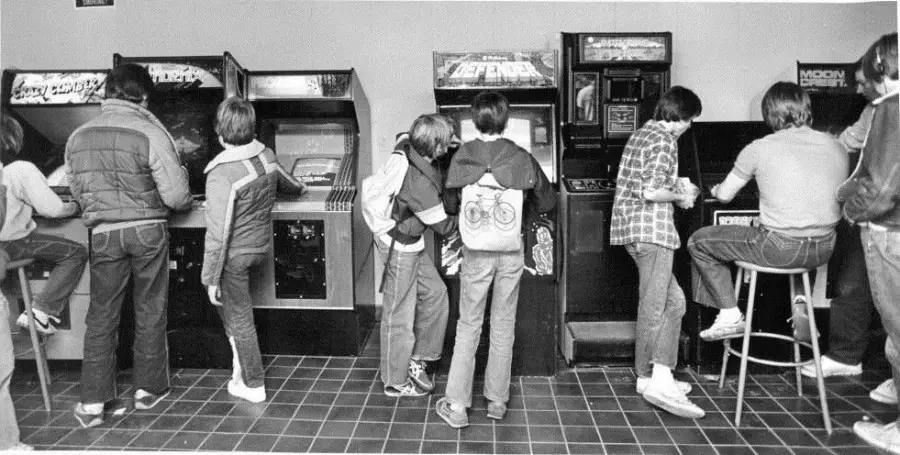 Black and white photo of kids in jackets and jeans playing arcade games, some standing and others sitting on stools, inside a retro arcade with tiled floors and various classic game machines.