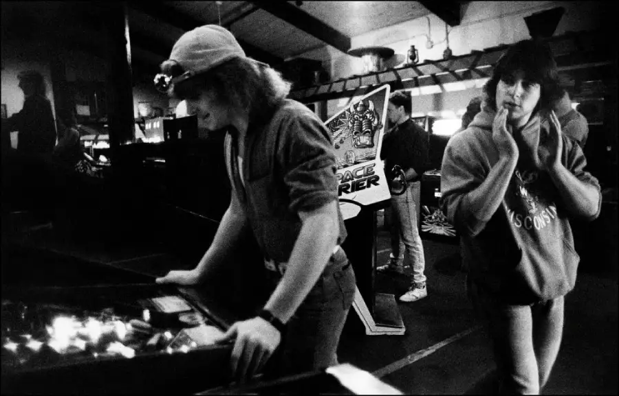 Two people stand by a pinball machine in a dimly lit arcade. One plays while the other watches. In the background, others stand near a Space Harrier arcade game. The atmosphere is casual and nostalgic.