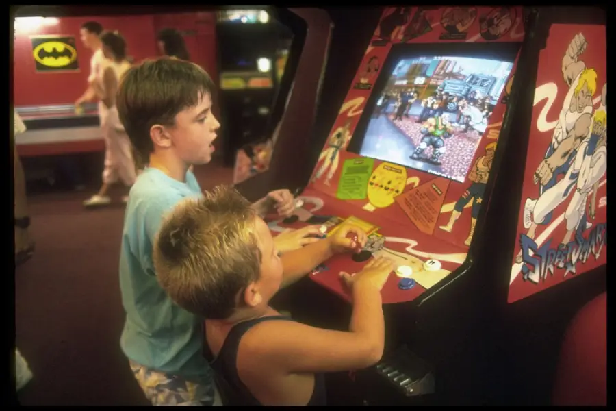 Two boys play a classic arcade fighting game on a colorful cabinet in an arcade. Other arcade machines and people are visible in the background.