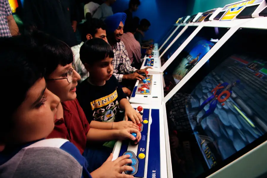 Children and adults play on arcade game machines, focused on the screens and controls. The scene is brightly lit, and the players are closely grouped together, enjoying the gaming experience.