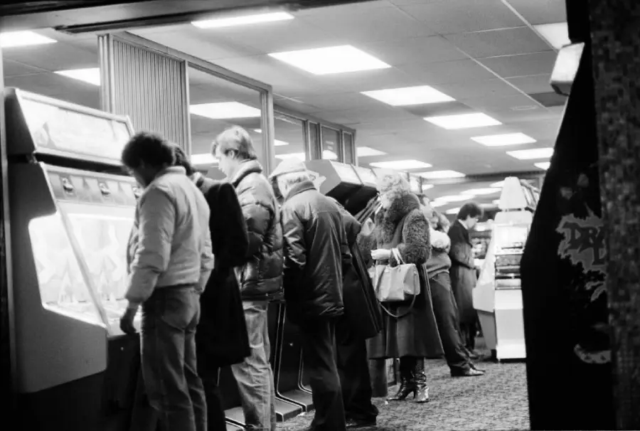 Black-and-white photo of people standing in a row, playing arcade machines in a brightly-lit gaming arcade. Most are wearing winter coats, and a few are carrying bags or purses.