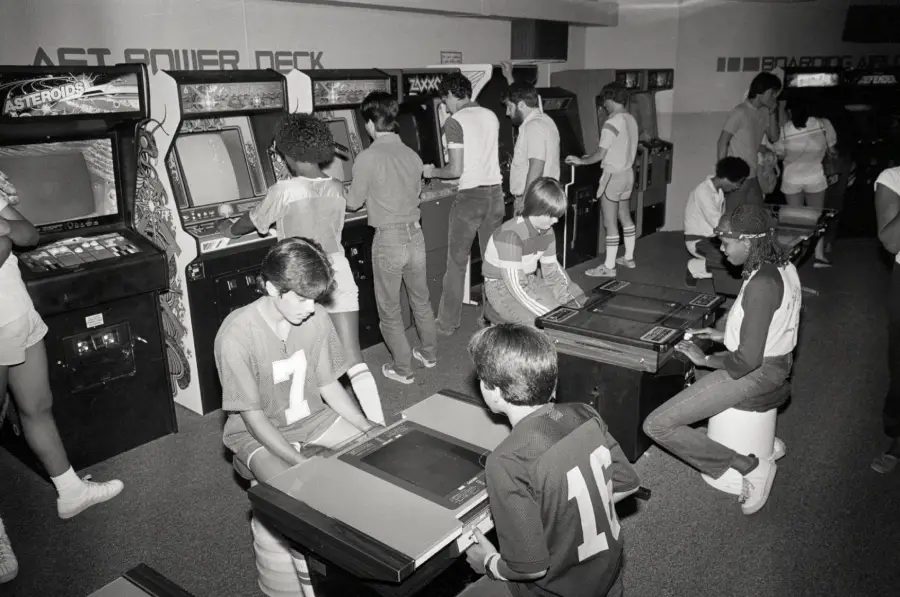 Black and white photo of a retro arcade. Teens play video games at upright and tabletop machines. Most wear jerseys, some sit, others stand, focused on screens. The arcade is busy and lively.