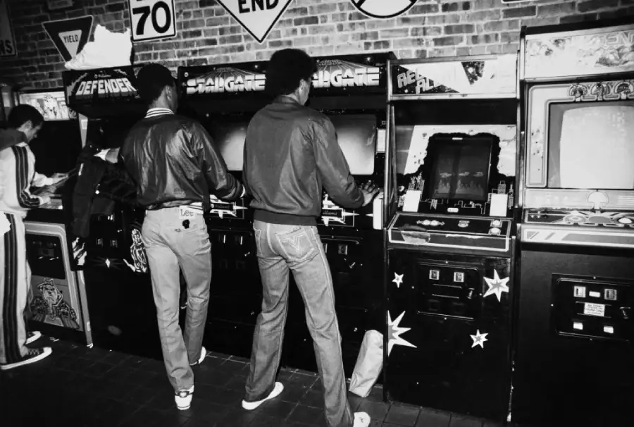 Two people stand at classic arcade game machines, playing games in a retro arcade. The scene features brick walls, vintage signs, and various arcade cabinets, creating a nostalgic atmosphere.