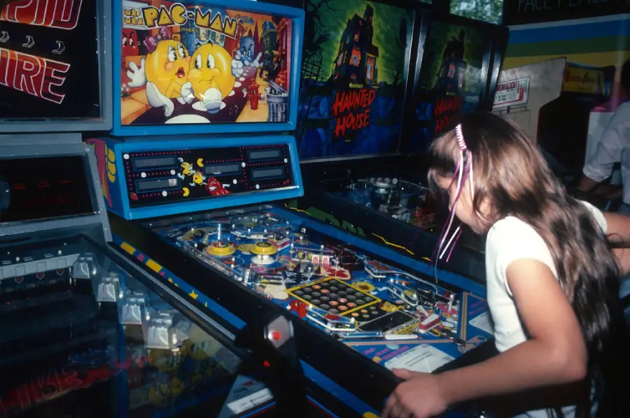 A young girl with long hair plays a pinball machine in an arcade, surrounded by other games, including machines featuring Pac-Man and Haunted House themes.