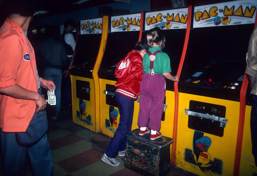 Two children play Pac-Man arcade machines, one standing on a box. An adult in an orange jacket holds cash nearby. The setting appears to be a dimly lit arcade.