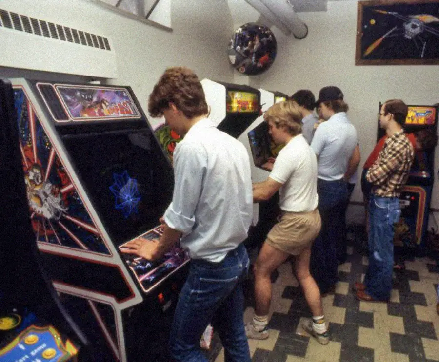 Five people play various arcade games in a retro arcade room with patterned flooring and posters on the walls. The players are focused on the screens of the classic arcade machines.