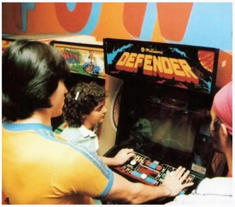 Three people play and watch a classic “Defender” arcade game in a retro arcade setting. The scene is colorful, with the game’s screen and controls visible, and the players appear focused and engaged.