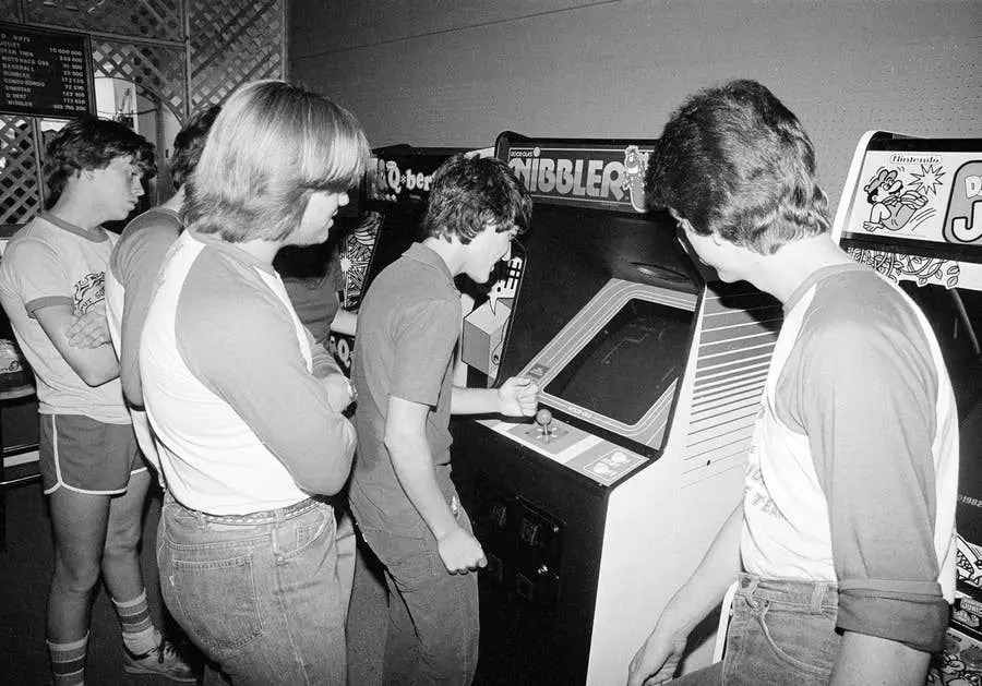 Five teenage boys play and watch at an arcade, focused on a “Nibbler” machine. The scene is in black and white, with the teens wearing casual 1980s clothing.