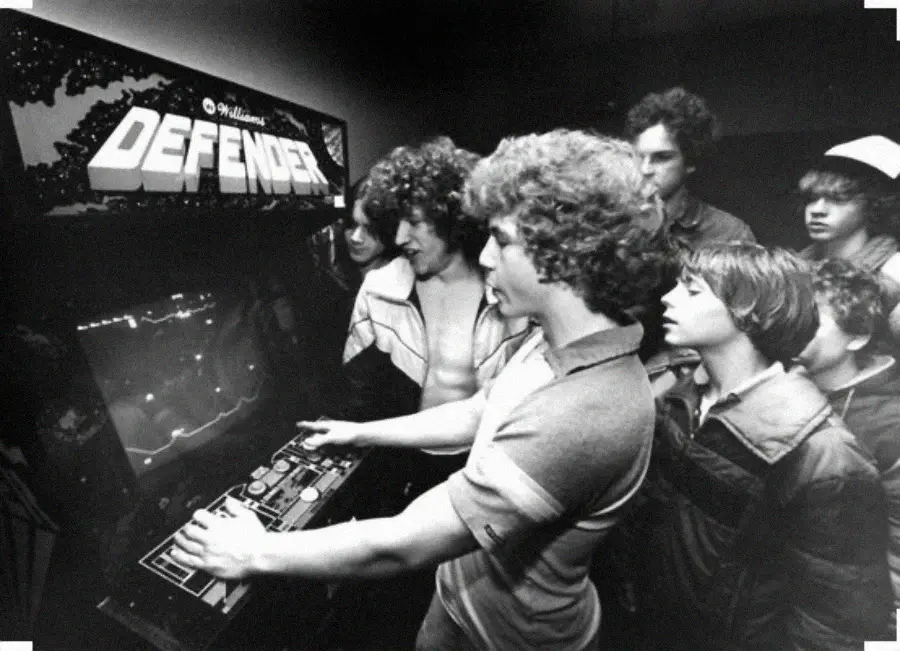 A group of boys gather around an arcade machine playing the game Defender, focused on the screen and controls in a dimly lit room.