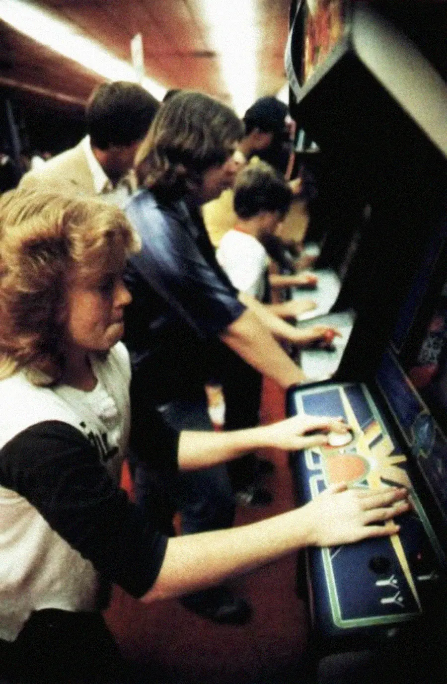 A group of young people play arcade games side by side in a dimly lit gaming arcade, focused on the game controls in front of them.