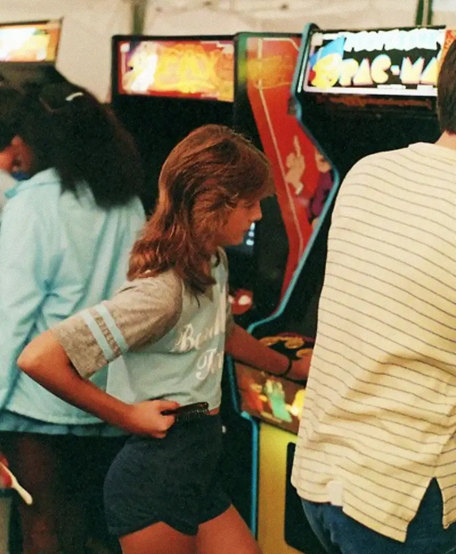A young person with a mullet hairstyle plays a classic Pac-Man arcade game in a busy arcade, surrounded by others and bright game machines.