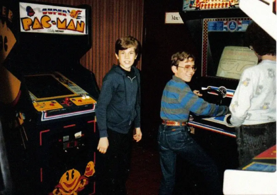 Three kids stand by vintage arcade machines, including PAC-MAN. One boy faces the camera, another plays a game, and the third stands nearby. The setting appears to be an old-school arcade.