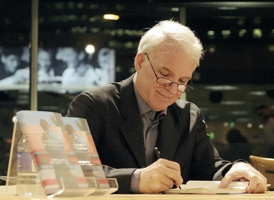 An older man with white hair and glasses is smiling while signing a book at a table. Books are stacked nearby, and he is indoors with blurred lights in the background.