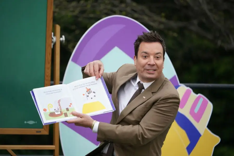 A man in a tan suit reads and shows a children’s picture book, smiling and gesturing animatedly. A colorful, egg-shaped decoration is visible in the background.