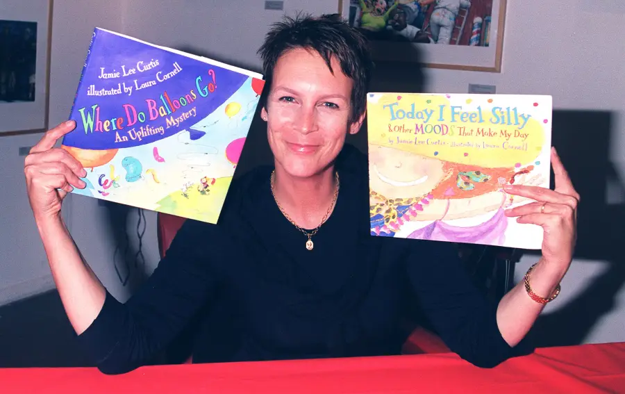 A woman smiles while holding up two children’s books: “Where Do Balloons Go?” and “Today I Feel Silly.” She sits at a red table with colorful art visible in the background.