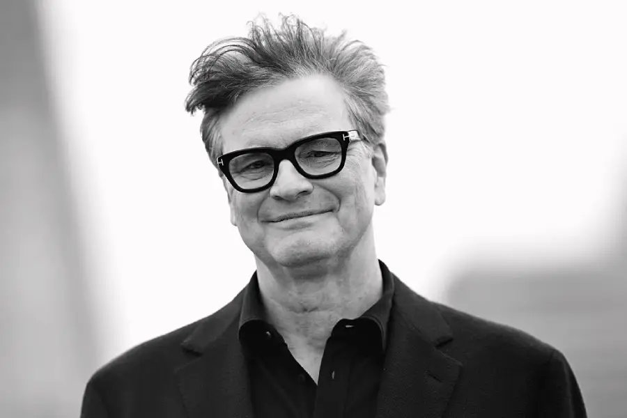 A man with glasses and tousled hair smiles slightly while wearing a dark jacket and shirt, standing outdoors with a blurred background.