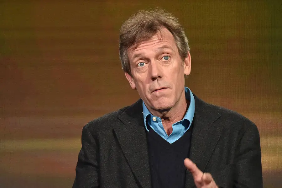 A man with light brown hair, wearing a blue shirt and black blazer, gestures with one hand while speaking against a blurred, warm-colored background.