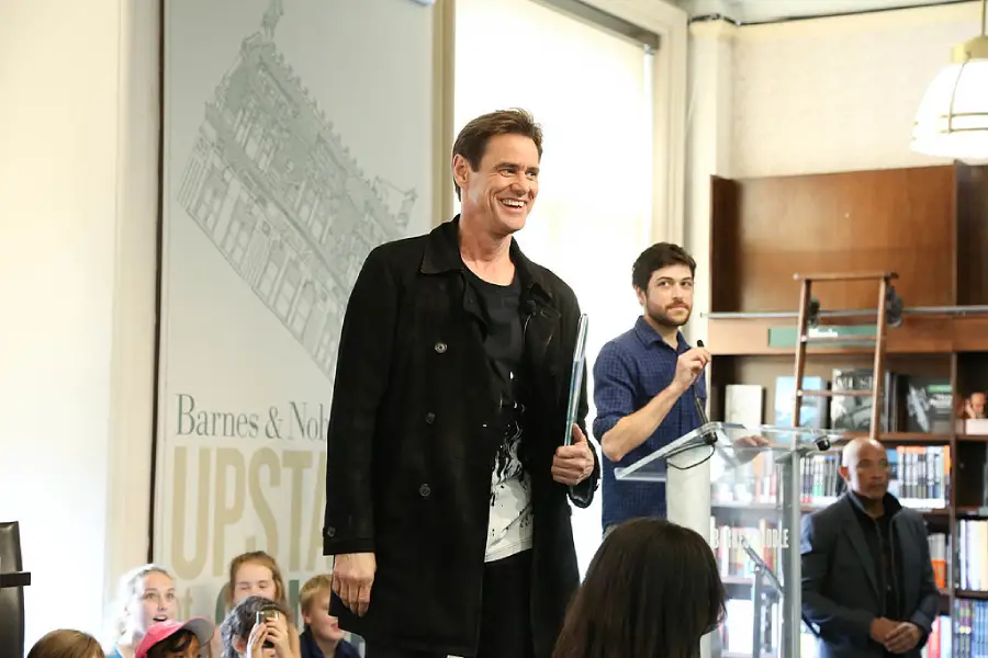 A man in a black coat smiles while standing in front of a seated audience at a bookstore event; another man stands at a podium with a microphone in the background.
