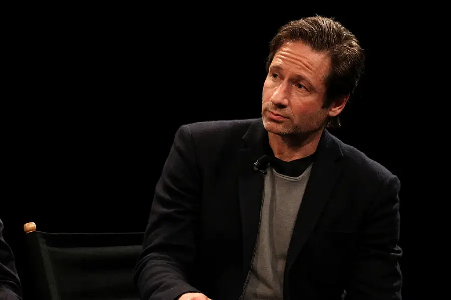 A man with short brown hair, wearing a dark blazer over a gray shirt, sits and looks thoughtfully to the side against a black background.