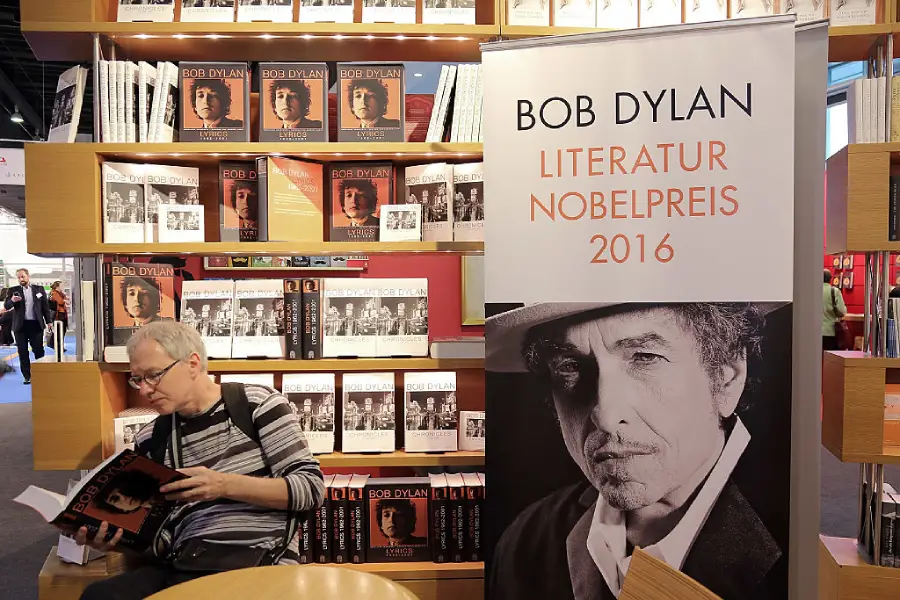 A person reads a Bob Dylan book in a bookstore, surrounded by shelves of the same book. A large banner celebrates Bob Dylan’s 2016 Nobel Prize in Literature.