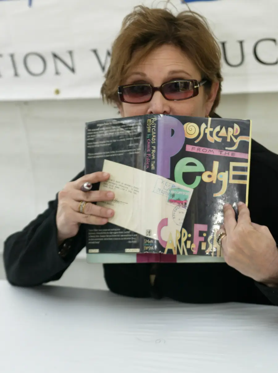 A person wearing sunglasses holds up the book “Postcards from the Edge” by Carrie Fisher, partially covering their face while seated at a table.