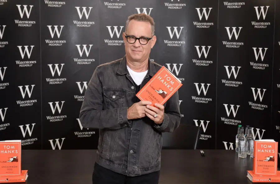 A man in a denim jacket holds a book titled “Tom Hanks: The Making of Another Major Motion Picture Masterpiece” at a Waterstones event, standing in front of a branded backdrop.