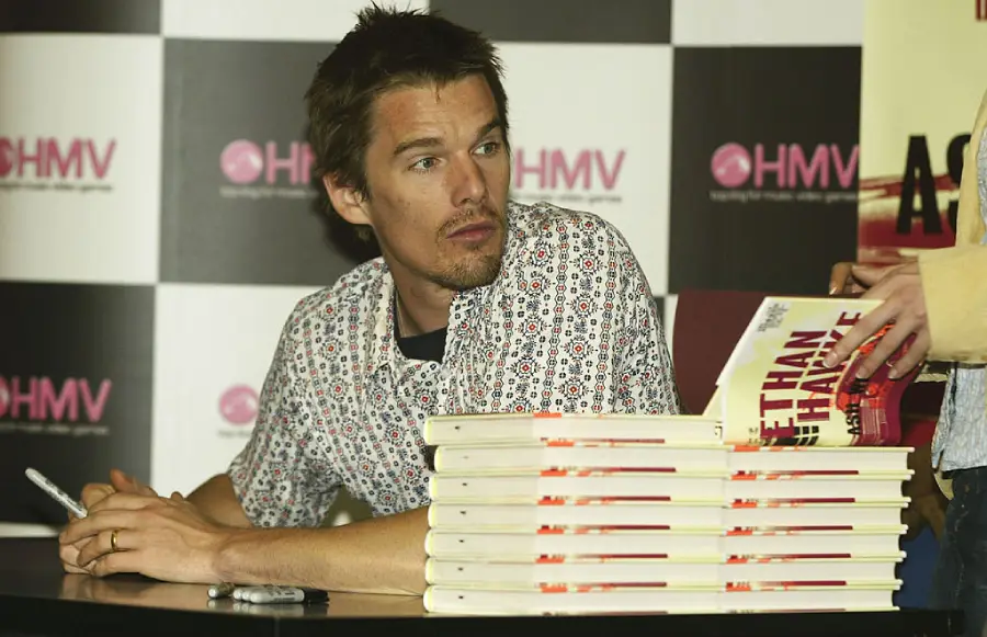 A man sits at a table with a stack of books, holding a pen, while another person hands him a book to sign. The backdrop features the HMV logo on a checkerboard pattern.