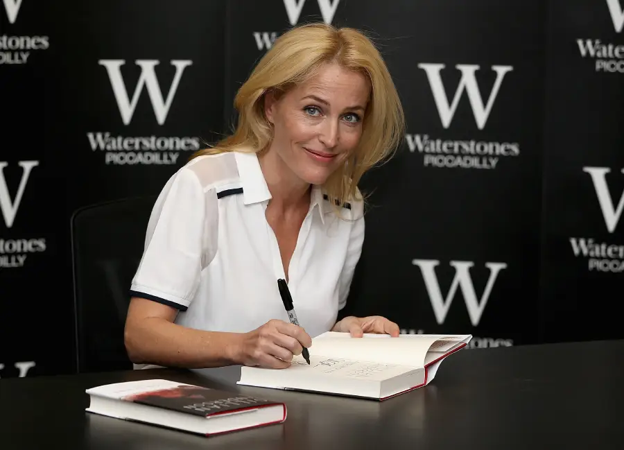 A woman with blonde hair smiles while signing a book at a table. She wears a white shirt, and another book sits beside her. Waterstones Piccadilly branding is visible on the black background behind her.