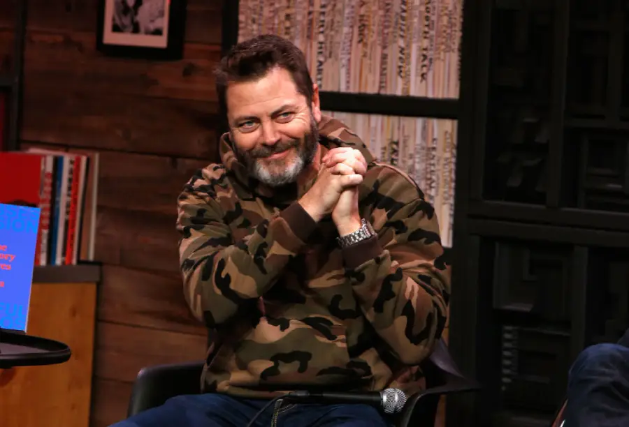 A bearded man in a camouflage hoodie sits and smiles with his hands clasped, in a cozy room with bookshelves and wood-paneled walls.