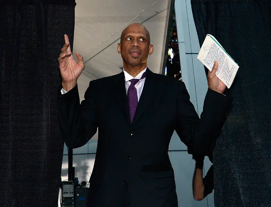 A man in a suit and tie stands between black curtains, holding a notepad in one hand and raising both hands, as if addressing an audience or preparing to speak.