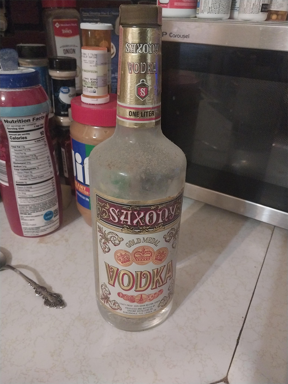A bottle of Saxony Vodka sits on a kitchen counter near peanut butter, spices, a can, and a microwave.