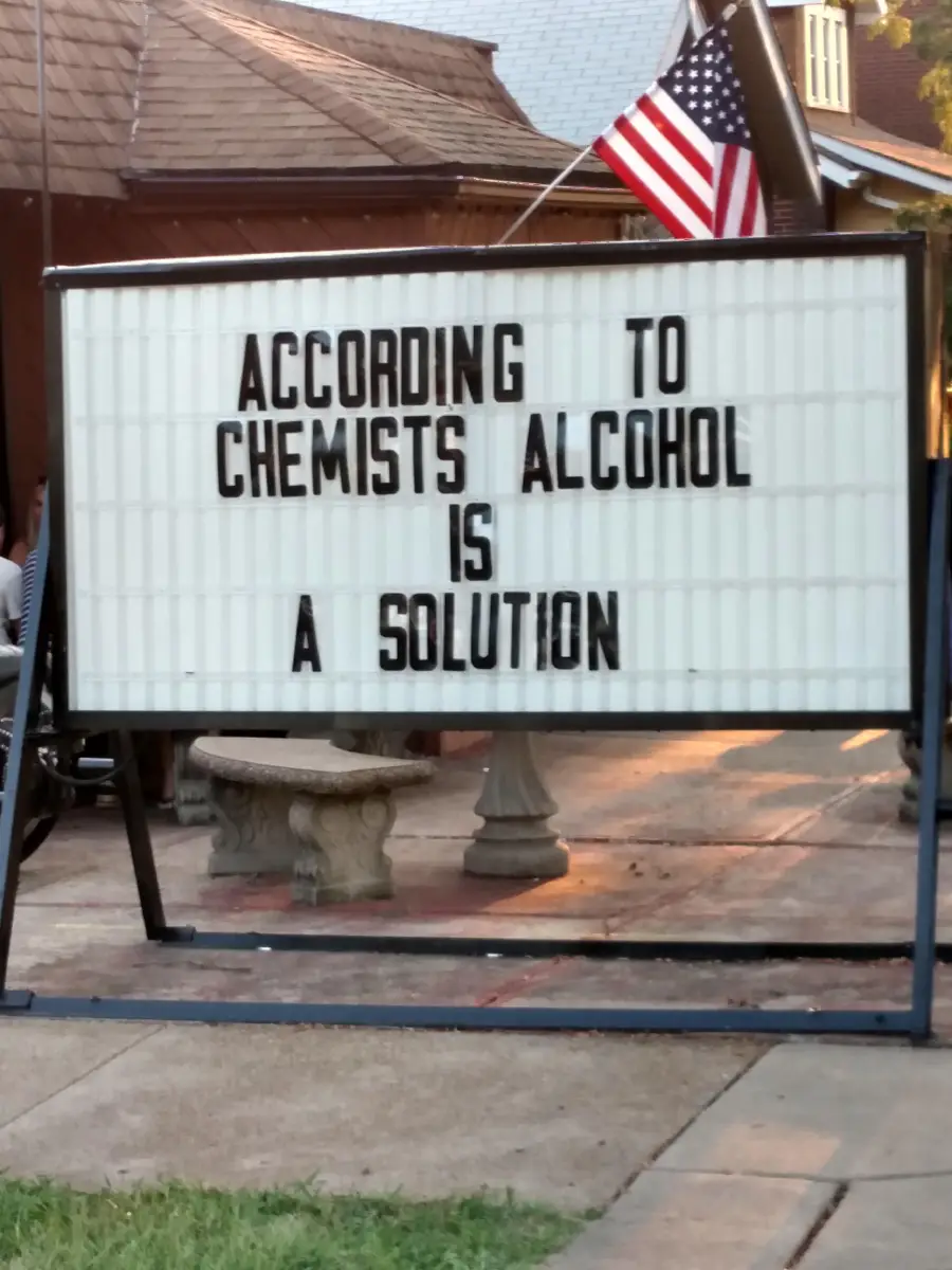 Outdoor sign reads: “According to chemists alcohol is a solution.” An American flag is above the sign and a stone bench is in the background.